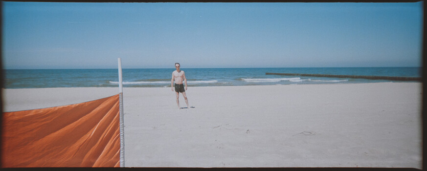 Person standing on wide sandy beach with volleyball net and orange fabric visible at frame edge.