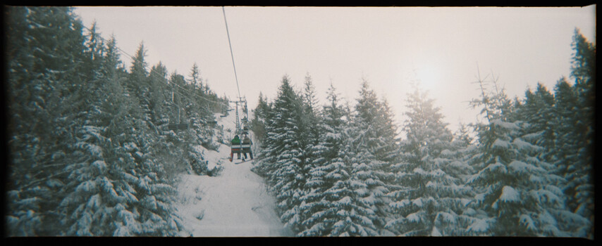Ski lift chairs suspended above a snow-covered forest of evergreen trees on an overcast day.