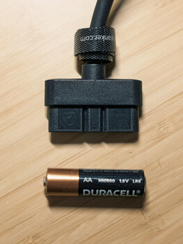 Camera flash battery pack and AA battery displayed on wooden surface for scale comparison.